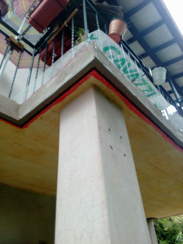 Kątowe ujęcie balkonu z widoczną kolumną wspierającą, zabezpieczoną siatką z nadrukiem, z donicami na balustradzie i świeżo pomalowanymi elementami na czerwono i brązowo.