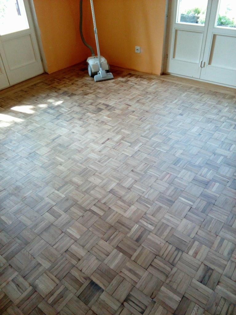 Parkiet ułożony w kwadraty, częściowo wyczyszczony, z odkurzaczem do parkietów w rogu pokoju, widoczne dwie pary drzwi.