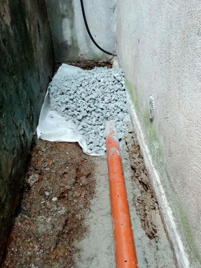 Drenaż fundamentów: rura drenarska z otworami, obsypana żwirem na geowłókninie, w wykopie przy ścianie budynku.