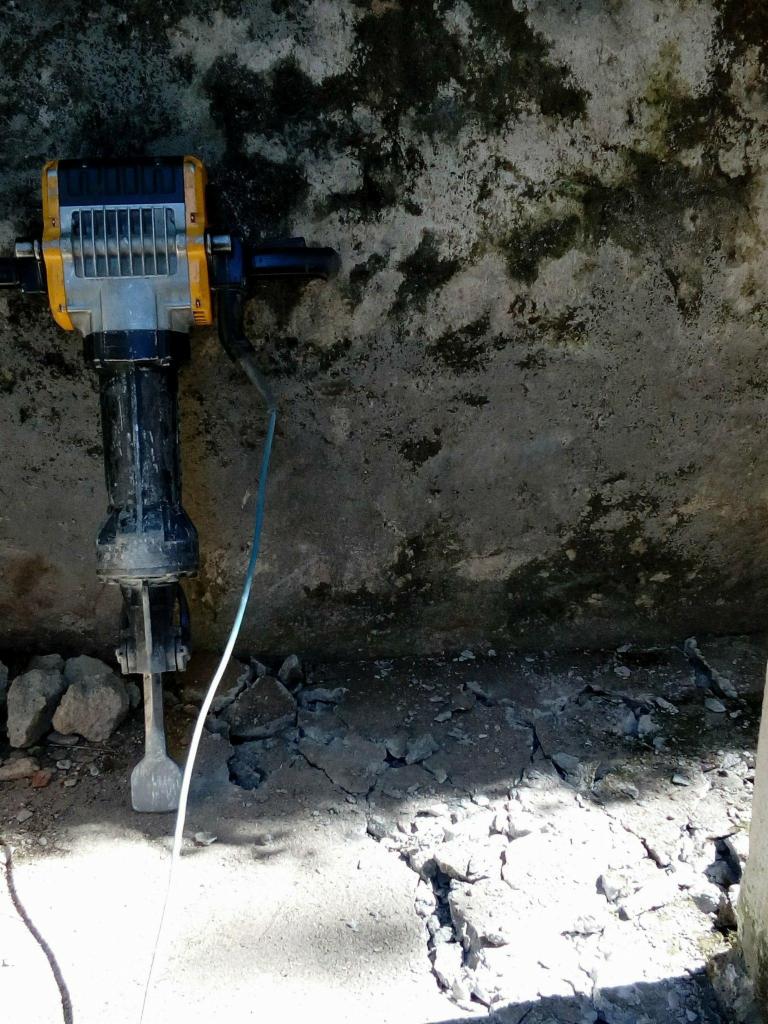 Żółto-czarny młot udarowy pneumatyczny oparty o szarą, wilgotną ścianę z fragmentami gruzu na podłożu.