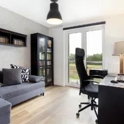 Nowoczesny pokój biurowy z szarą sofą z wzorzystymi poduszkami, czarną szafką z książkami, biurkiem z komputerem i lampą oraz oknem z widokiem na zieleń.
