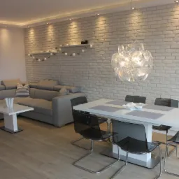 Jasne wnętrze: szara sofa narożna z poduszkami, biały stolik kawowy, biały stół z przezroczystymi krzesłami, dekoracyjna ściana z białej cegły z półkami i girlandą świetlną, designerska lampa wisząca.