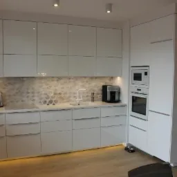 Nowoczesna, biała kuchnia z połyskującymi frontami szafek, mozaikową ścianą nad blatem i oświetleniem LED pod szafkami. Widoczne urządzenia AGD: ekspres do kawy, mikrofalówka i piekarnik.