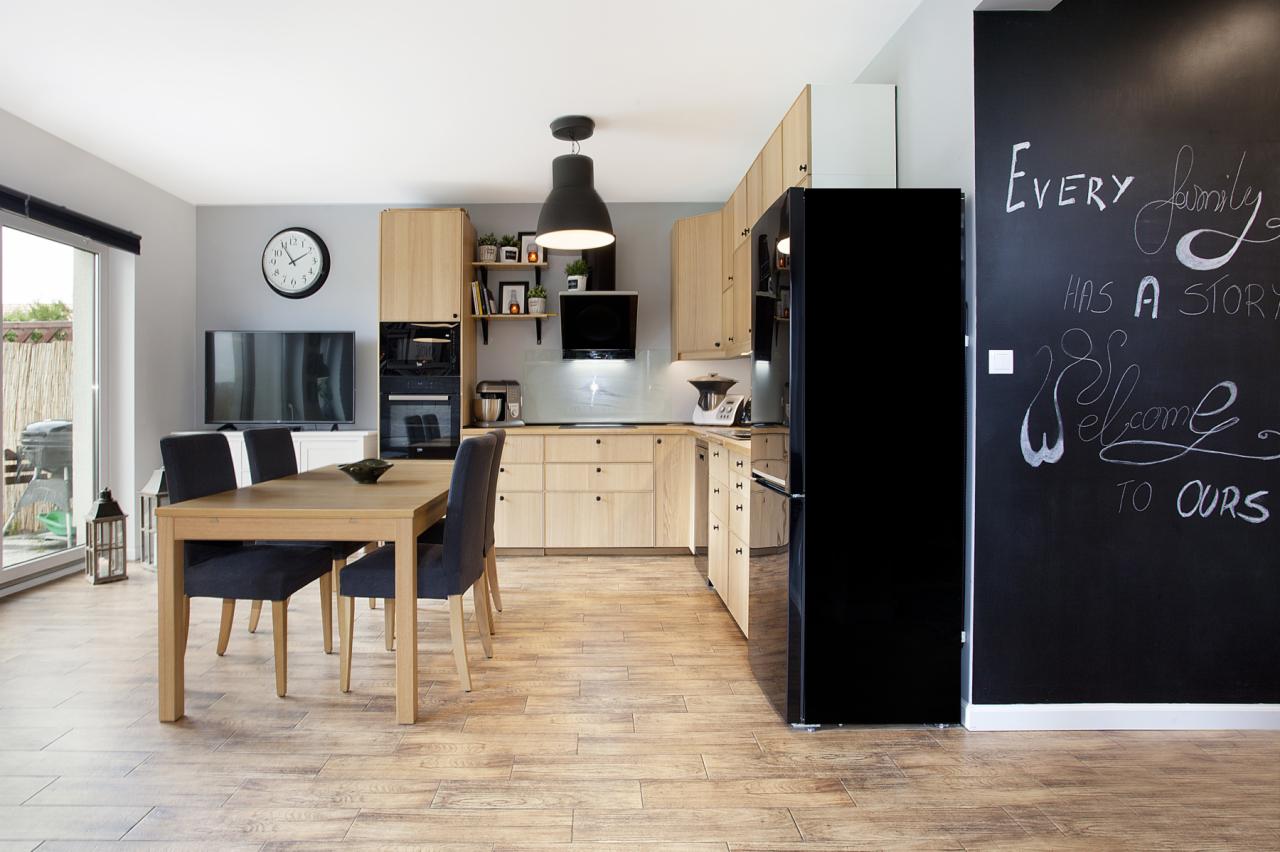 Jasna kuchnia z drewnianymi meblami, czarną lodówką i ścianą z napisem kredą, widok na stół jadalny i okno balkonowe.