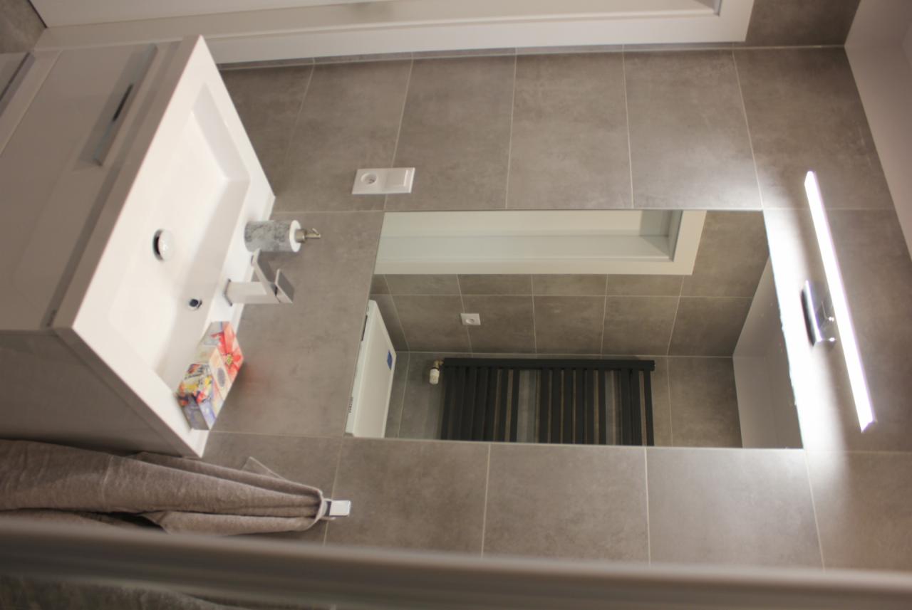 Nowoczesna łazienka z szarymi płytkami, białą umywalką z szafką, minimalistyczną baterią, lustrem z podświetleniem LED i grzejnikiem drabinkowym widocznym w odbiciu lustra.