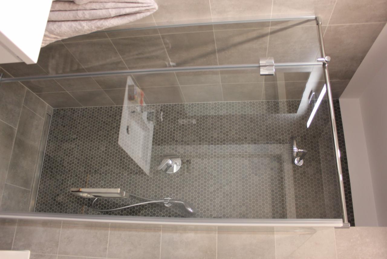 Nowoczesna kabina prysznicowa z szarą mozaiką heksagonalną na podłodze, szklane drzwi z chromowanymi zawiasami, widok z góry.