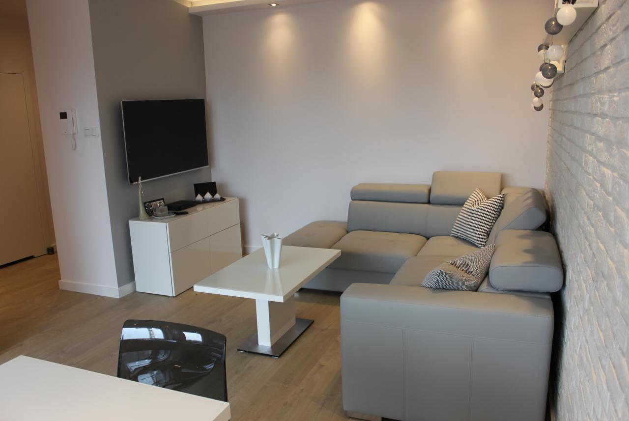 Nowoczesny salon z narożną, szarą sofą z regulowanymi zagłówkami, białą ścianą z cegły, białym stolikiem kawowym z wazonem i szafką RTV pod telewizorem.