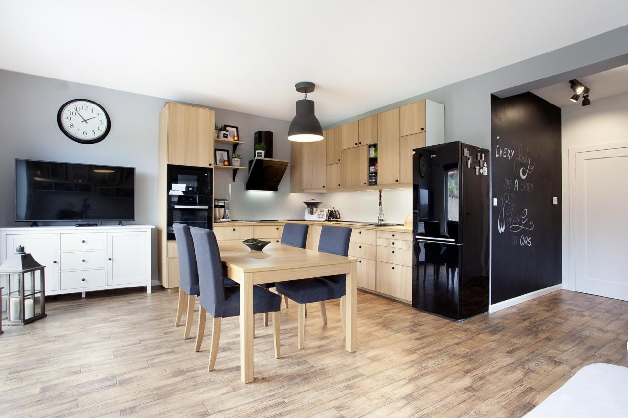 Jasna kuchnia z drewnianymi frontami, czarną lodówką i ścianą pomalowaną farbą tablicową z białymi napisami, stół z czterema krzesłami.