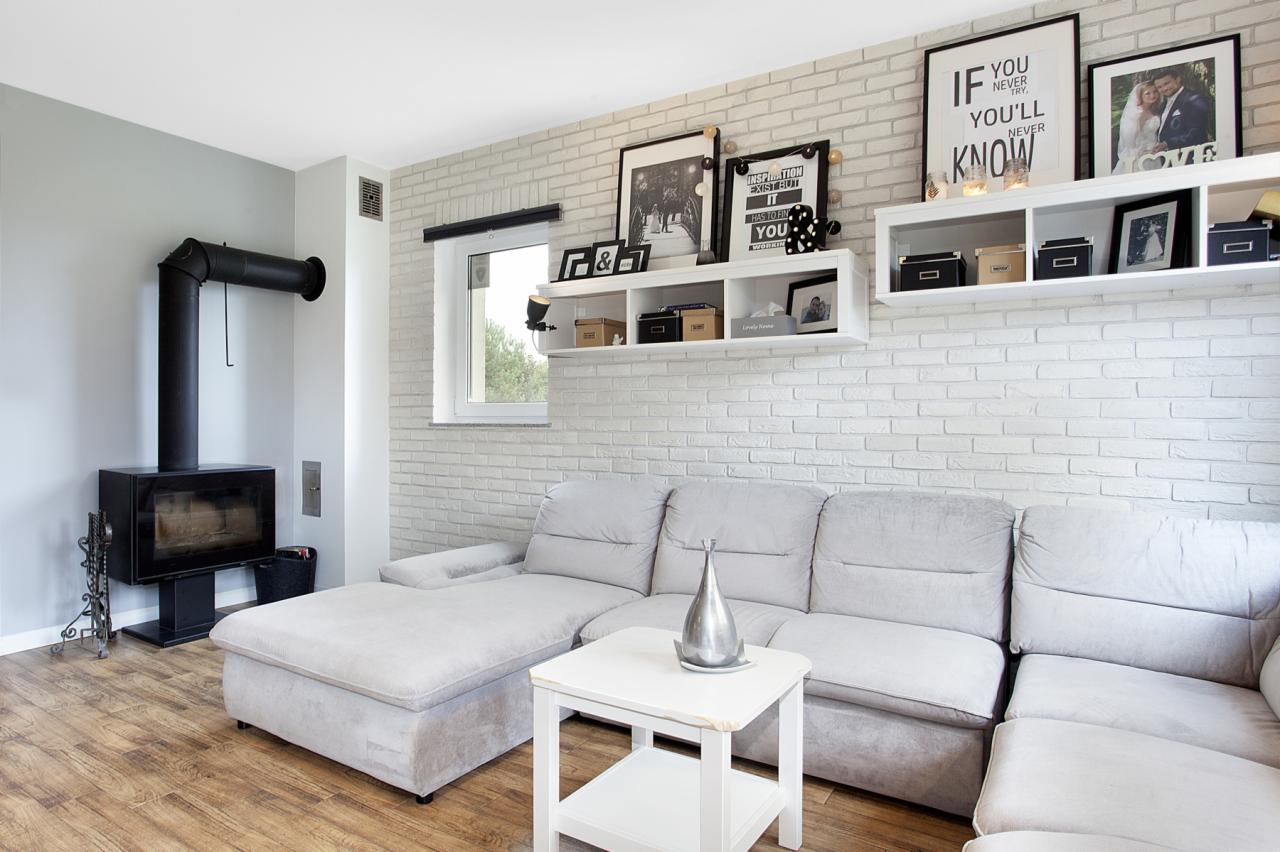 Jasnoszary narożnik z szezlongiem, biały stolik kawowy i kominek w salonie z białą ceglaną ścianą, dekoracje na półkach i drewniana podłoga.