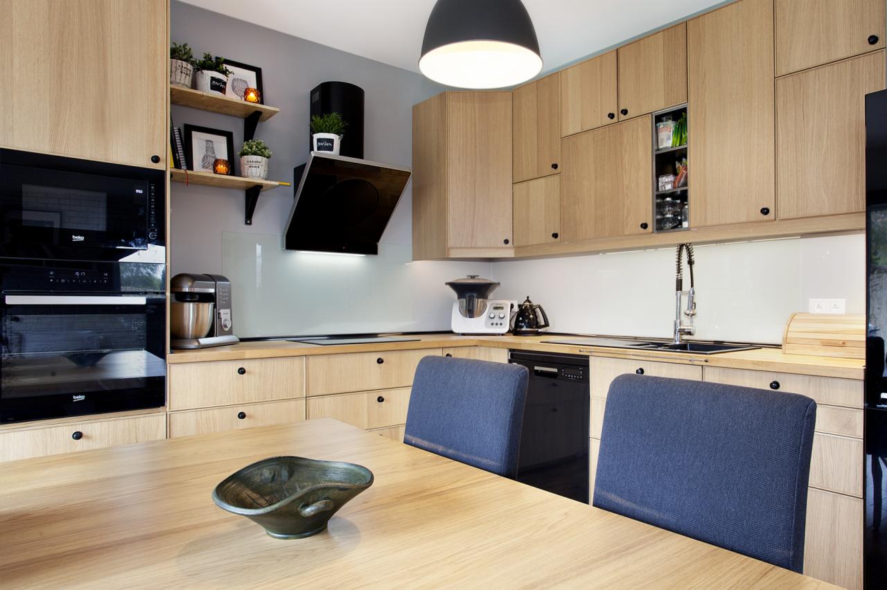 Nowoczesna kuchnia z drewnianymi szafkami, czarnymi akcentami i minimalistycznym oświetleniem. Na pierwszym planie drewniany stół z dekoracyjną misą i dwoma niebieskimi krzesłami.