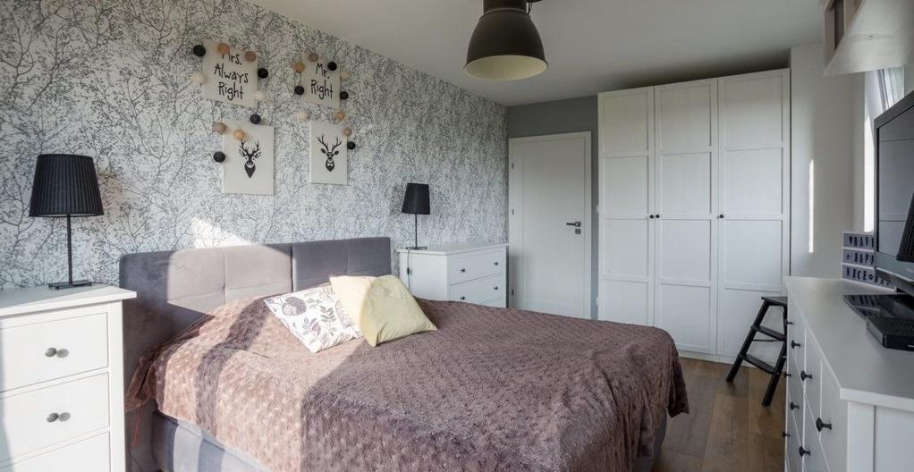Sypialnia z łóżkiem z szarym zagłówkiem, brązową narzutą, białymi komodami, szafą w zabudowie, tapetą w motywy gałązek i dekoracjami ściennymi z napisami 'Mrs. Always Right' i 'Mr. Right'.