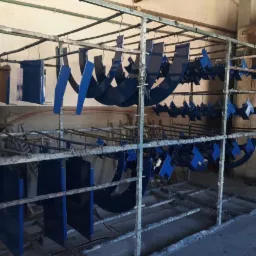 Metalowe elementy konstrukcyjne, świeżo po aplikacji niebieskiej farby, wiszą na wieszakach w metalowej ramie, gotowe do wyschnięcia w hali.