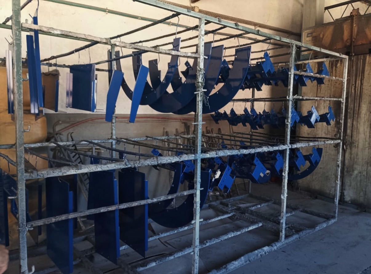 Metalowe elementy konstrukcyjne, świeżo po aplikacji niebieskiej farby, wiszą na wieszakach w metalowej ramie, gotowe do wyschnięcia w hali.