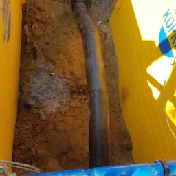 Czarna rura kanalizacyjna w wykopie zabezpieczonym żółtymi panelami, połączona z niebieską rurą wodociągową. W tle widoczna ziemia i niewielka kałuża.