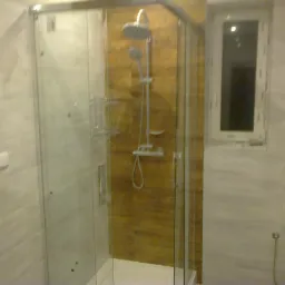 Kabina prysznicowa z przezroczystymi drzwiami, widoczna armatura i brązowe płytki wewnątrz, na ścianach białe płytki imitujące marmur, obok grzejnik drabinkowy i okno.