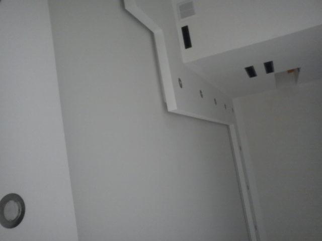 Nowoczesne wnętrze z białą ścianą, sufitem podwieszanym i oświetleniem punktowym, widoczne otwory wentylacyjne i instalacyjne.