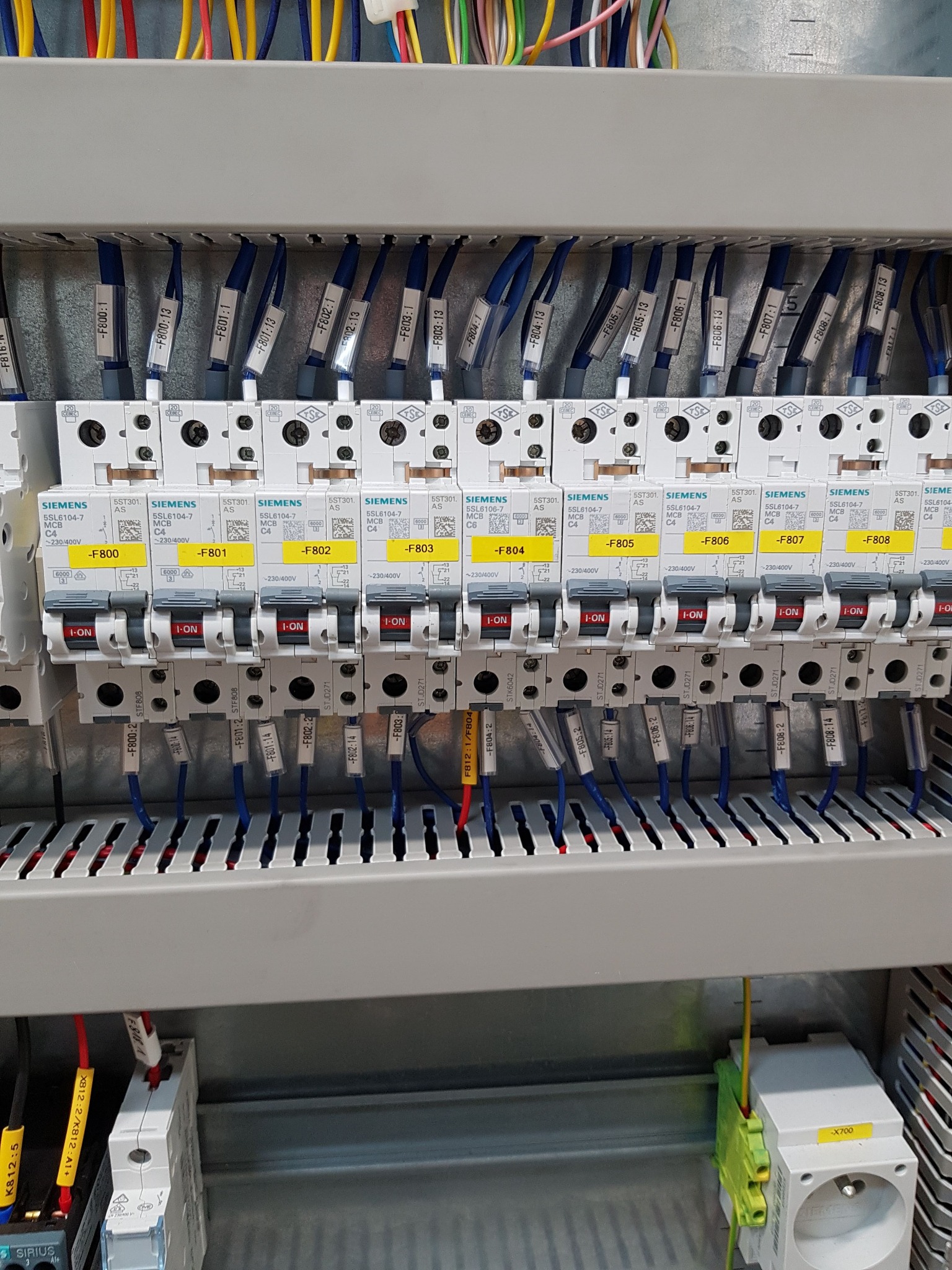 Panel sterowania z zamontowanymi bezpiecznikami Siemens, oznaczonymi etykietami, z widocznymi niebieskimi przewodami i żółtymi naklejkami identyfikacyjnymi.
