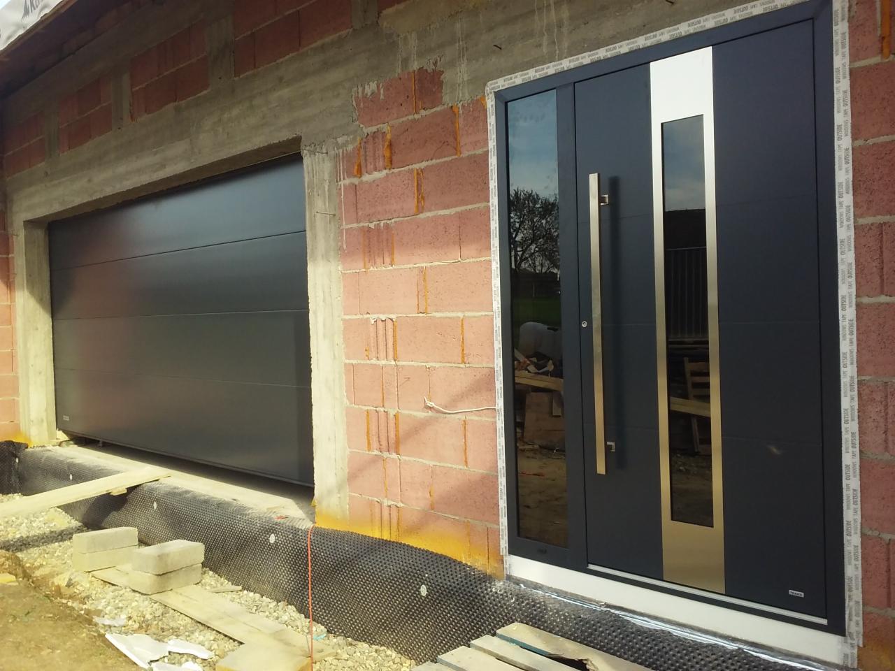 Nowoczesne, antracytowe drzwi wejściowe z przeszkleniem i pionowym, srebrnym panelem, obok brama garażowa w trakcie montażu, widoczne materiały budowlane i surowa cegła.