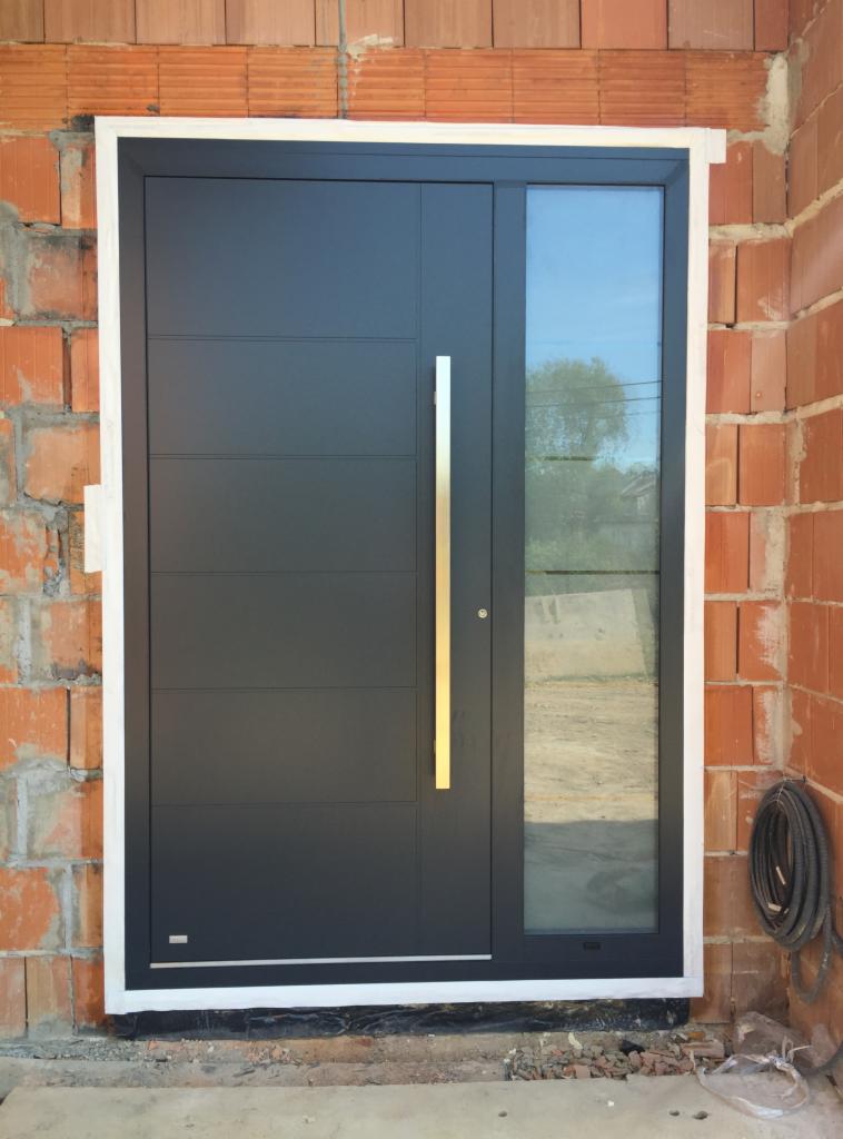 Nowoczesne, antracytowe drzwi zewnętrzne z długim, pionowym uchwytem i bocznym przeszkleniem, osadzone w surowym murze z czerwonej cegły.