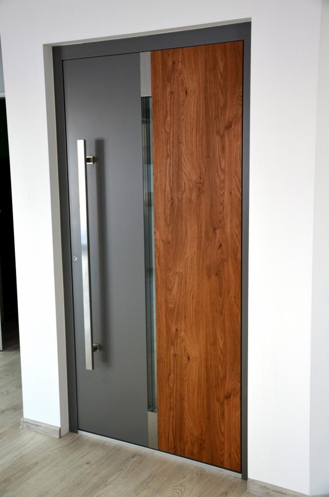 Nowoczesne drzwi wejściowe z szarą ramą, długim, srebrnym uchwytem i drewnianym panelem bocznym o wyraźnym usłojeniu.