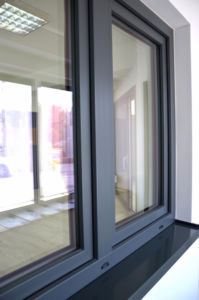 Szczegółowe ujęcie szarego okna aluminiowego z widocznym parapetem i odbiciem wnętrza pomieszczenia, ukazującym podłogę i fragmenty innych okien.