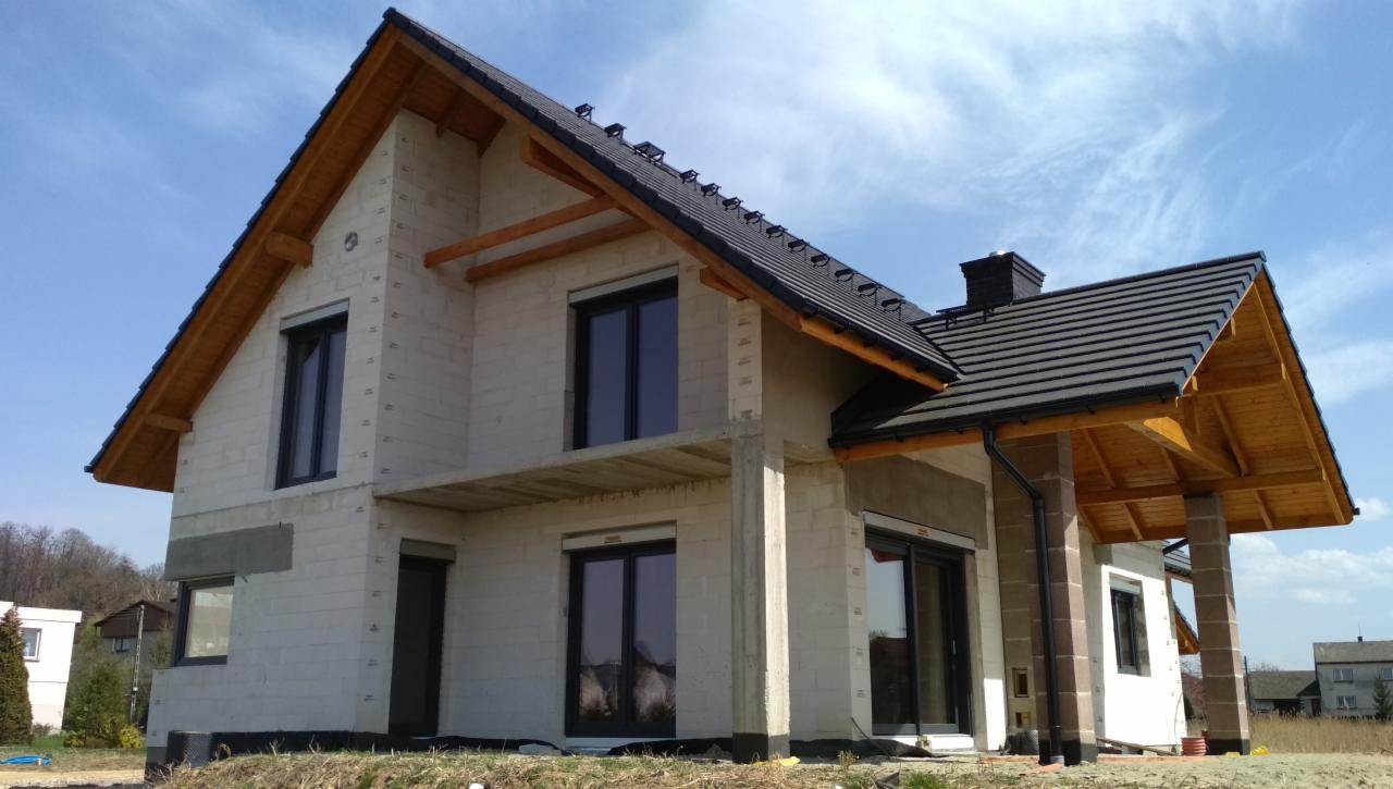 Dom w trakcie budowy z zamontowanymi ciemnymi oknami i częściowo położonym dachem z drewnianą podbitką, widoczne elementy konstrukcyjne budynku.