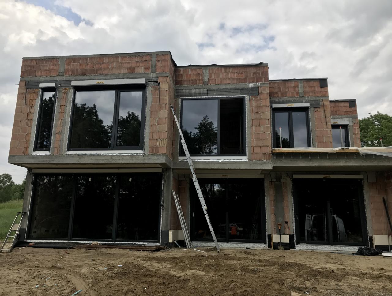 Surowy budynek w trakcie budowy z zamontowanymi, dużymi, czarnymi oknami i częściowo opuszczonymi roletami zewnętrznymi. Widoczne drabiny i narzędzia budowlane.