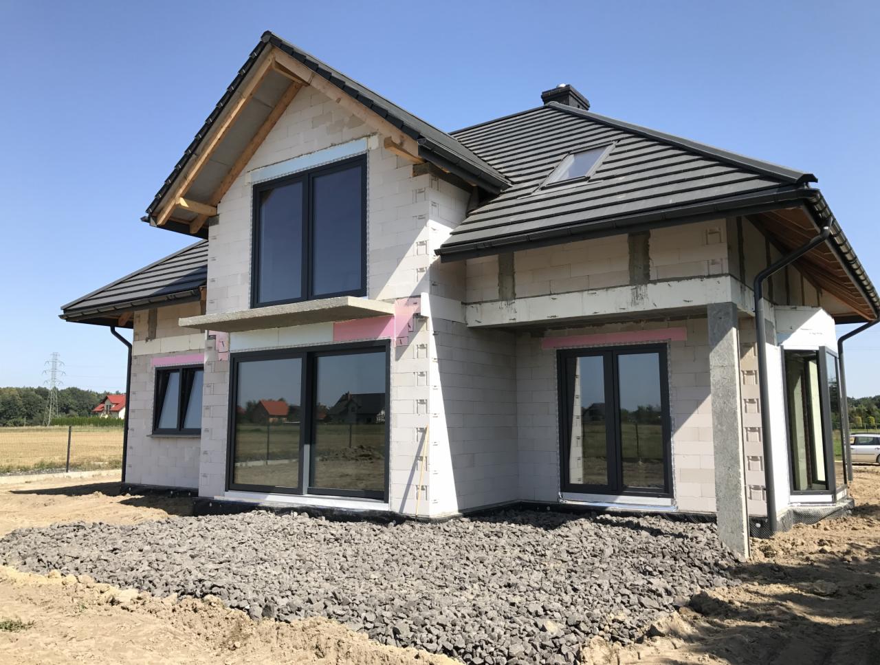 Dom w trakcie budowy z zamontowanymi oknami aluminiowymi w grafitowych ramach, widoczne ocieplenie wokół okien, teren wokół domu częściowo wysypany kamieniem.