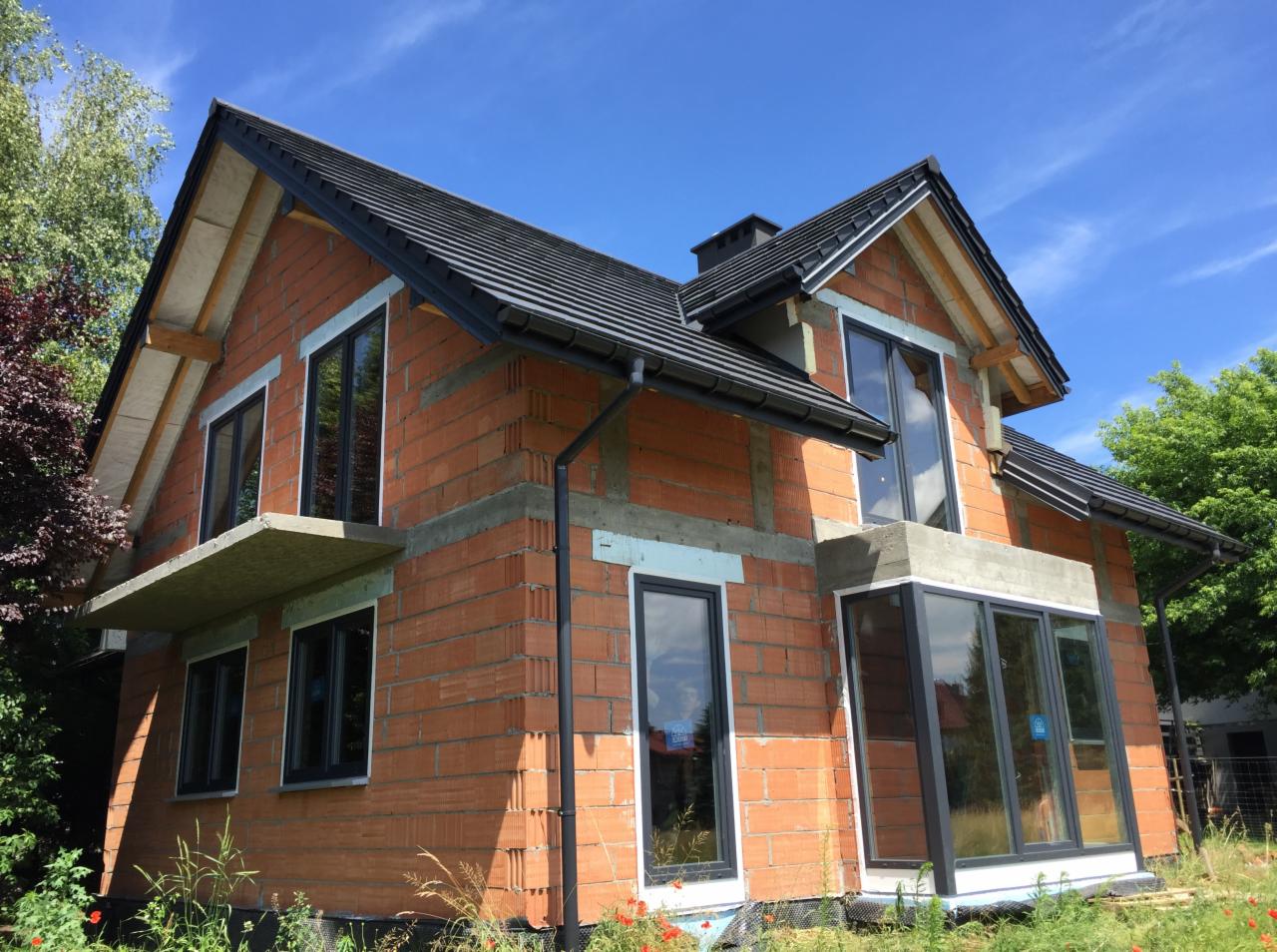 Dom w trakcie budowy z czerwonej cegły, czarne ramy okienne, szara dachówka, widoczne ocieplenie styropianem, błękitne niebo w tle.