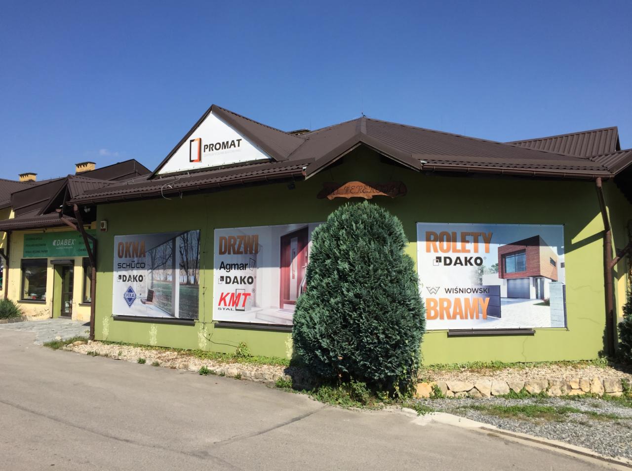 Budynek firmy Promat z reklamami okien, drzwi i rolet firm Schüco, Agmar, Dako i Wiśniowski, zasłonięty częściowo przez krzew.
