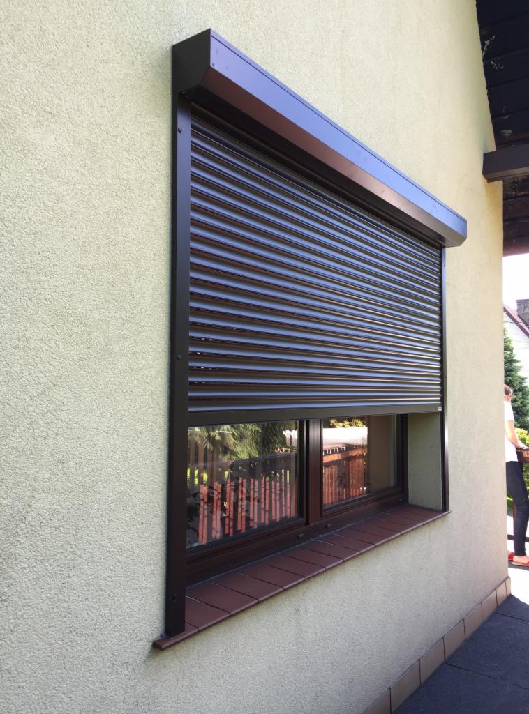 Brązowa roleta zewnętrzna całkowicie zasłonięta na oknie budynku z teksturowaną, beżową elewacją i parapetem wyłożonym brązowymi płytkami.
