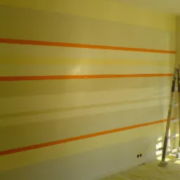 Ściana w pokoju pomalowana w poziome pasy w odcieniach żółci i pomarańczy, z drabina malarską opartą o ścianę i folią malarską na podłodze.