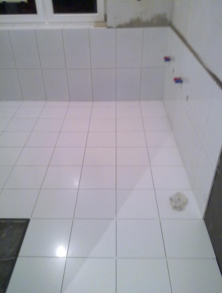 Wnętrze łazienki w trakcie kładzenia białych płytek, widoczne wystające przyłącza wodne z czerwonymi i niebieskimi zaślepkami, resztki zaprawy na ścianie i szmatka na podłodze.