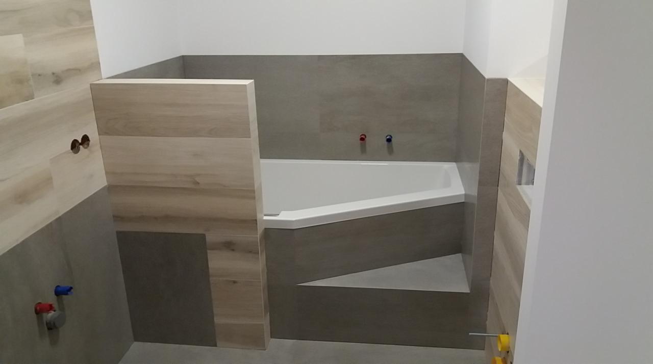 Wykończona łazienka z narożną wanną obudowaną szarymi płytkami, z elementami wykończenia imitującymi drewno oraz widocznymi przyłączami wodnymi z czerwonym i niebieskim zaworem.