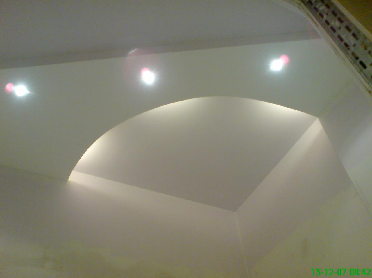 Biały sufit z łukowatym obniżeniem i wbudowanymi punktowymi źródłami światła, widoczne fragmenty instalacji elektrycznej przy krawędzi sufitu.