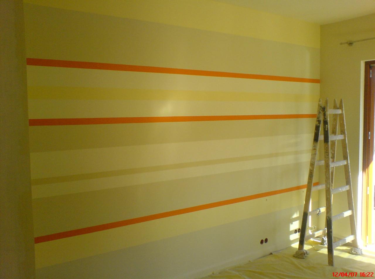 Ściana w pokoju pomalowana w poziome pasy w odcieniach żółci i pomarańczy, z drabina malarską opartą o ścianę i folią malarską na podłodze.