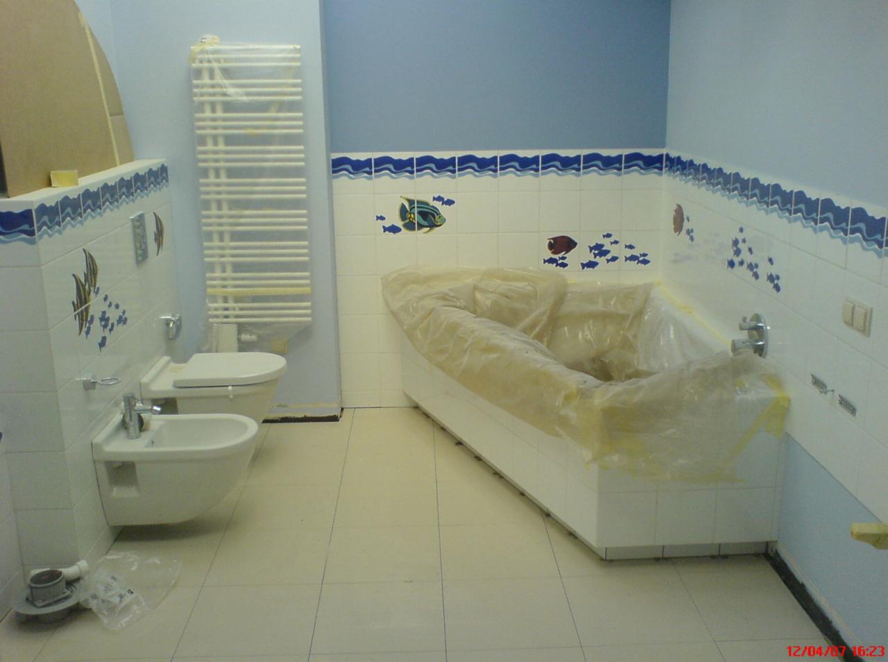 Łazienka w trakcie zabezpieczania przed pracami wykończeniowymi: wanna owinięta folią ochronną, ściany wyłożone białymi płytkami z motywem ryb, grzejnik drabinkowy zabezpieczony folią, widoczne...