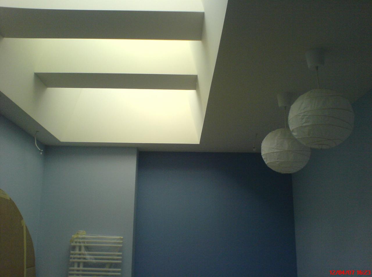 Wnętrze pokoju z niebieskimi ścianami, białym grzejnikiem, dwoma papierowymi lampionami pod sufitem i nietypowym oknem dachowym z wnękami.