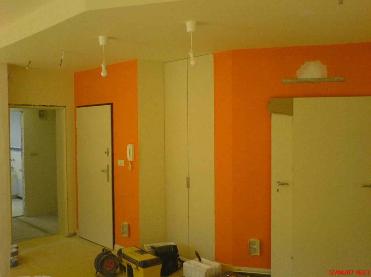 Pokój po częściowym malowaniu ścian na pomarańczowo, widoczne narzędzia malarskie, niezabezpieczone gniazdka i niedomalowane miejsce na ścianie.