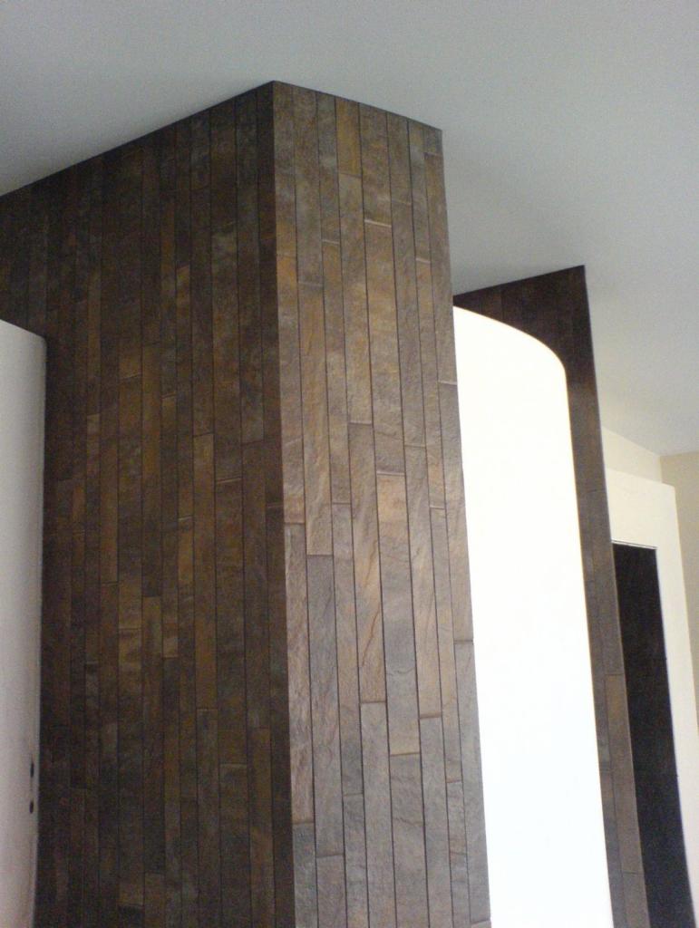 Dekoracyjna ściana wyłożona wąskimi, pionowymi płytkami imitującymi kamień w odcieniach brązu i szarości, przylegająca do białej ściany i sufitu.