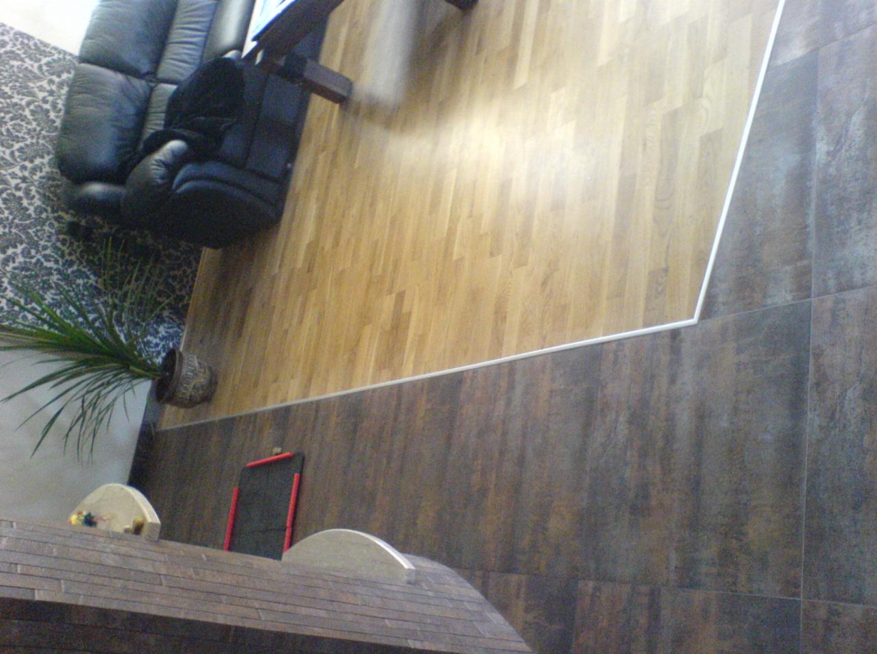 Połączenie paneli podłogowych z brązowymi płytkami, widok z góry na fragment salonu z czarną sofą i rośliną doniczkową.