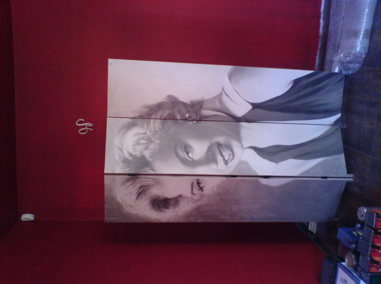 Trzyczęściowy obraz z wizerunkiem Marilyn Monroe zawieszony na czerwonej ścianie, widoczne niedoskonałości montażu i fragment podłogi z zabawkami.