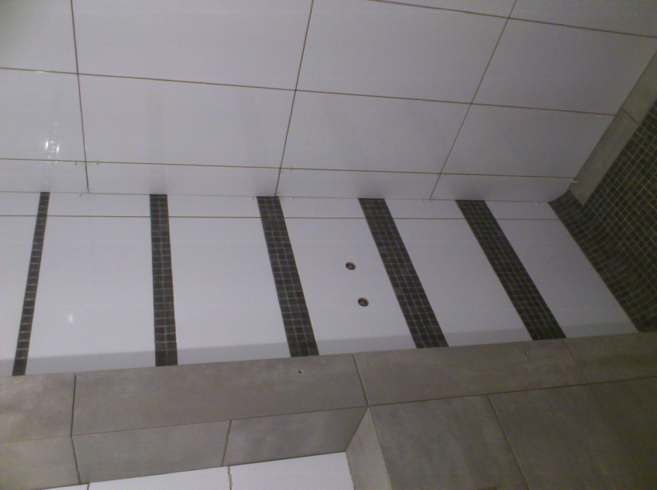 Fragment wykończonej łazienki z białymi płytkami na ścianach i podłodze, ułożonymi w regularny wzór, z ciemnymi paskami mozaiki wkomponowanymi w podłogę. Widoczne dwa otwory w podłodze...