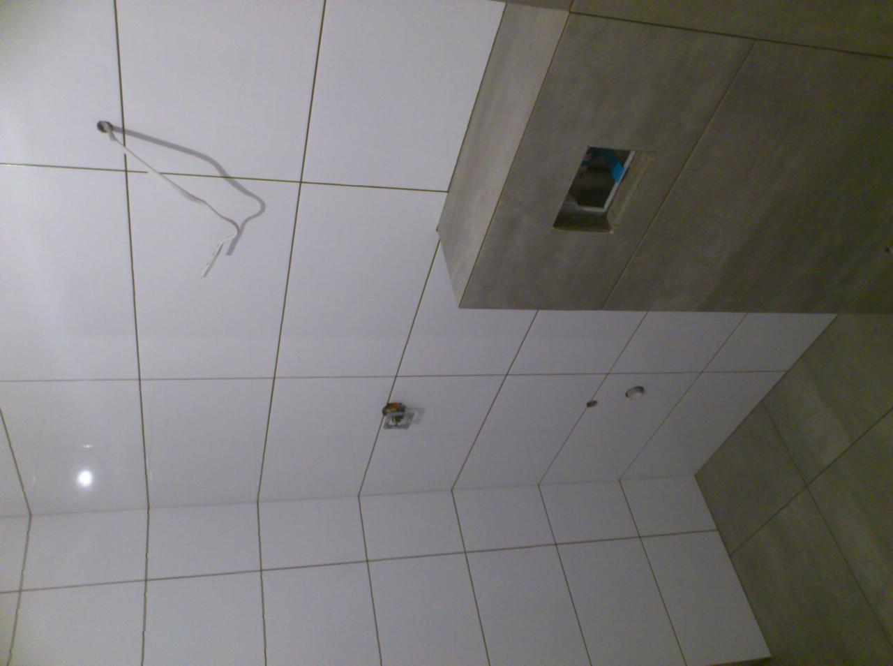 Białe płytki na ścianie i suficie łazienki w trakcie wykańczania, widoczne wystające kable i otwór na oświetlenie.