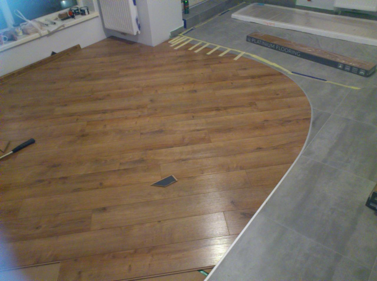 W trakcie układania paneli podłogowych w pomieszczeniu o nietypowym kształcie, widoczne docięte panele i narzędzia, przejście do podłogi z płytek zaznaczone taśmą maskującą.