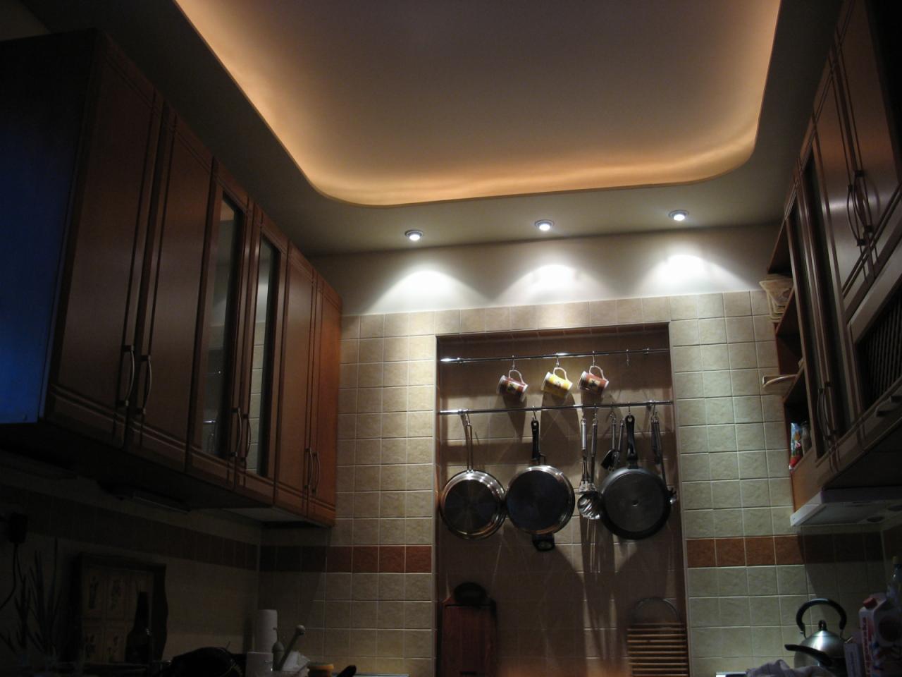 Kuchnia z brązowymi szafkami, podwieszanym sufitem z oświetleniem LED, ceramiczną mozaiką na ścianach oraz wiszącymi patelniami i kubkami.