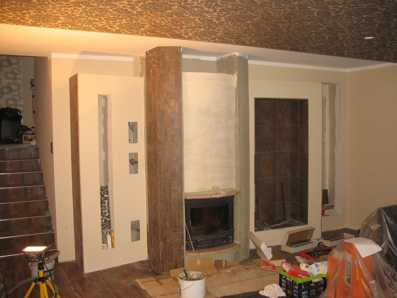 Wnętrze w trakcie prac wykończeniowych: obudowa kominka z okładziną imitującą drewno, ściany w beżowym odcieniu, widoczne narzędzia i materiały budowlane, dekoracyjny sufit.