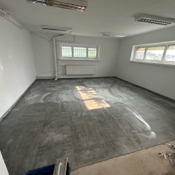 BUD-ART - Szare płytki podłogowe po czyszczeniu w pustym, odnowionym pokoju z oknami i grzejnikiem, widoczne narzędzia glazurnicze.