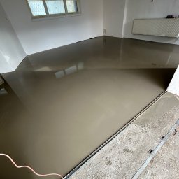 BUD-ART - Wylewka samopoziomująca na podłodze w jasnym pomieszczeniu, widoczne kable i listwa dylatacyjna przy ścianie.