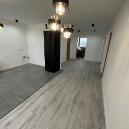 Wnętrze mieszkania w trakcie prac wykończeniowych: szare płytki podłogowe przechodzą w panele podłogowe imitujące drewno, białe ściany, czarna lodówka i nowoczesne lampy wiszące.